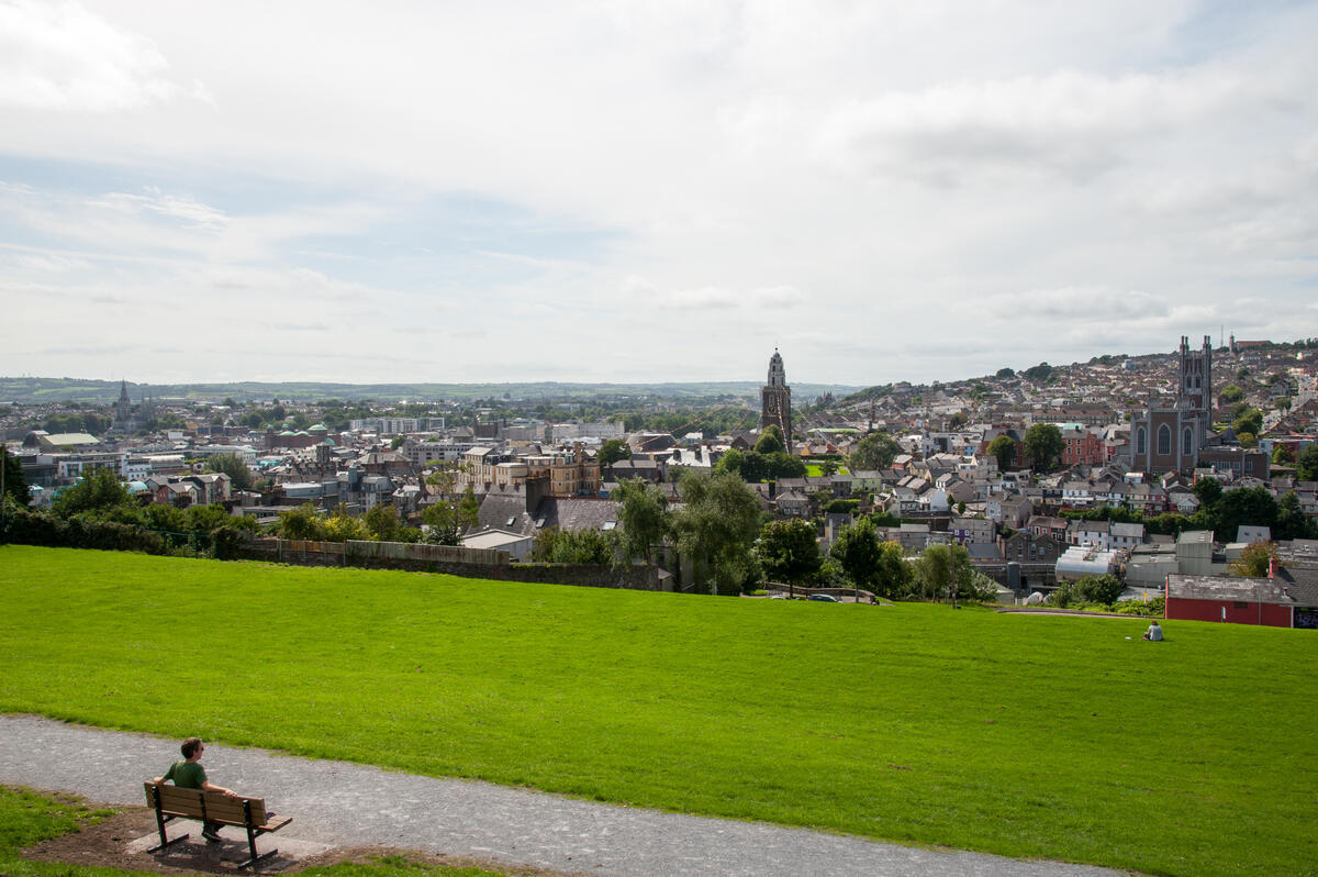 A wide view across Cork City from Patrick&rsquo;s Hill, with green foreground, church towers, rooftops and distant countryside.